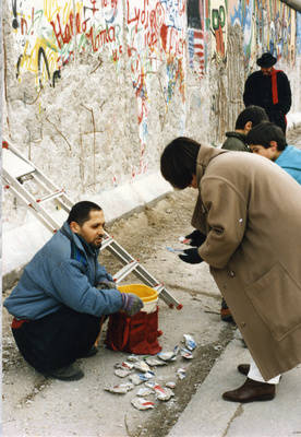 Ein Mauerstück-Händler kniet an der Grenzmauer 75 vor einer Sammlung Mauerstücke und bietet sie Passanten an
