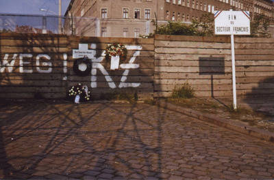 Mit Kränzen geschmücktes Weißes Kreuz an der Betonschichtmauer Bernauer Straße / Eberswalder Straße